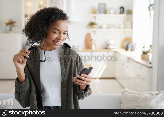 Smiling young mixed race girl using mobile apps, holding smartphone, shopping in online store at home. Modern teen lady with afro hairstyle chatting in social network, reading message with good news.. Smiling young mixed race girl using mobile apps, holding phone, shopping in online store at home
