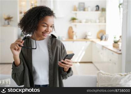 Smiling young mixed race girl using mobile apps, holding smartphone, shopping in online store at home. Modern teen lady with afro hairstyle chatting in social network, reading message with good news.. Smiling young mixed race girl using mobile apps, holding phone, shopping in online store at home
