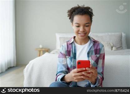 Smiling young mixed race girl using wireless earphones, mobile apps on smartphone at home, talking online by video call. Modern biracial teen lady chatting, listening to music, sitting in bedroom.. Smiling mixed race girl using earphones, phone apps, talking online by video call sitting in bedroom