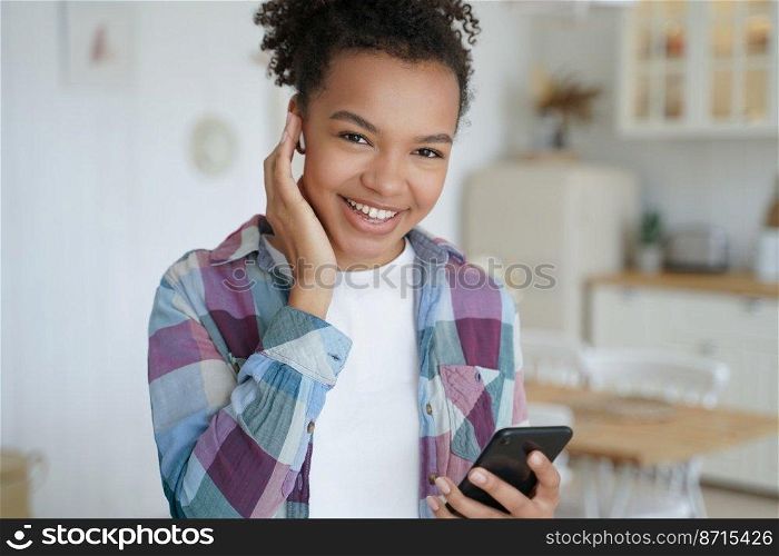 Smiling young mixed race teen girl listens to music in wireless earphone holding smartphone, using mobile apps at home. Happy biracial teen lady enjoy sound, listening to audio book or podcast indoors. Smiling mixed race girl listens to music in earphone holding smartphone, using mobile apps at home
