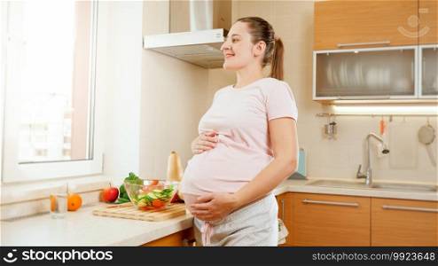 Smiling young pregnant woman touching and holding big abdomen looking out of window on big kitchen.Concept of healthy lifestyle and nutrition during pregnancy.. Smiling young pregnant woman touching and holding big abdomen looking out of window on big kitchen.Concept of healthy lifestyle and nutrition during pregnancy