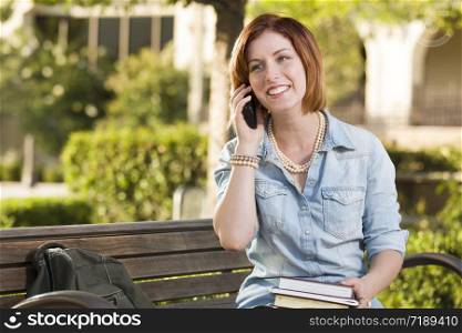 Smiling Young Pretty Female Student Outside on Cell Phone with Backpack and Books Sitting on Bench.
