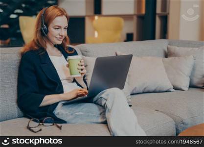 Smiling young redhaired woman chatting with friends online via laptop and wireless headset earphones, while sitting on sofa in modern coffee house interior, holding cup with drink in her hand. Smiling young red haired woman chatting with friends online via laptop and wireless headset earphones