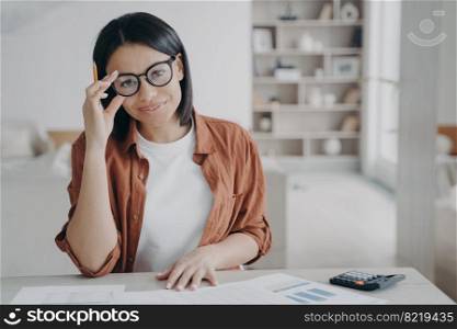 Smiling young woman accountant freelancer wearing glasses looking at camera sitting at desk at home. Female bookkeeper working with financial statistics of company, makes financial audit.. Smiling female accountant freelancer in glasses working looking at camera sitting at desk at home