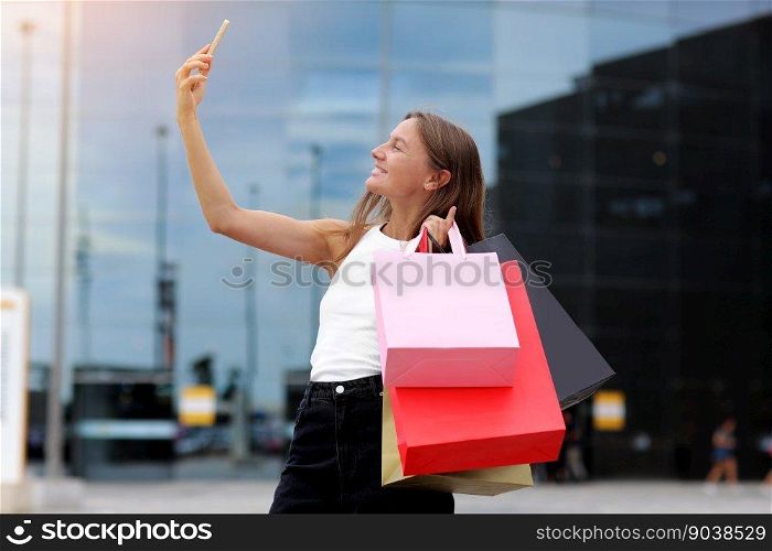 Smiling young woman blogger with shopping bags and smartphone is communicating with followers in social networks near shopping mall. concept of consumerism, sale, rich life, virtual life.Black Friday.. Smiling young woman blogger with shopping bags and smartphone is communicating with followers in social networks near shopping mall. concept of consumerism, sale, rich life, virtual life. Black Friday