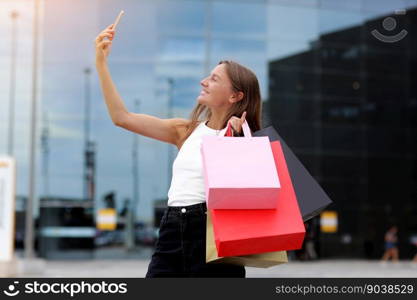 Smiling young woman blogger with shopping bags and smartphone is communicating with followers in social networks near shopping mall. concept of consumerism, sale, rich life, virtual life.Black Friday.. Smiling young woman blogger with shopping bags and smartphone is communicating with followers in social networks near shopping mall. concept of consumerism, sale, rich life, virtual life. Black Friday