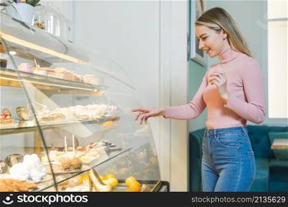 smiling young woman choosing cake standing front glass cabinet