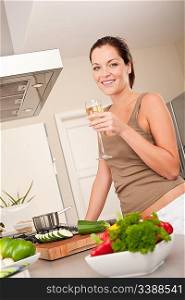 Smiling young woman cooking and holding glass of white wine in the kitchen