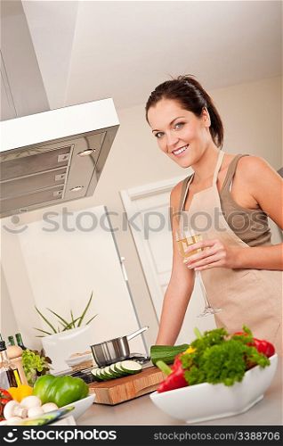 Smiling young woman cooking and holding glass of white wine in the kitchen