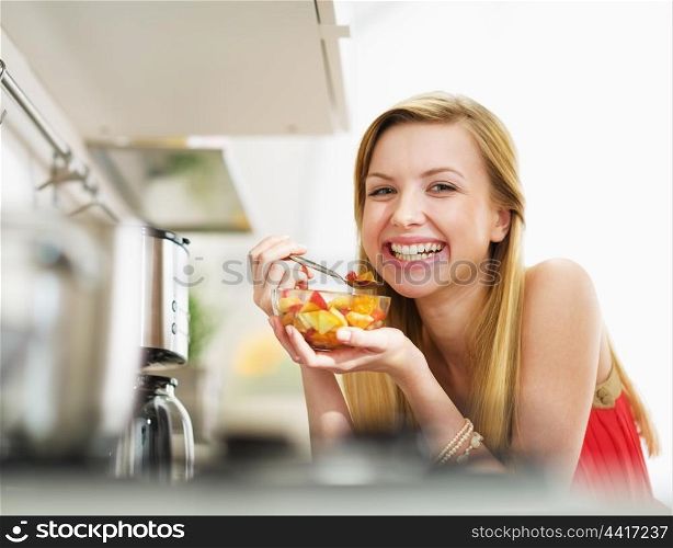 Smiling young woman eating fresh fruits salad in kitchen