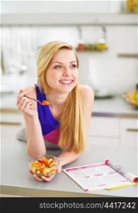 Smiling young woman eating fruits salad in kitchen