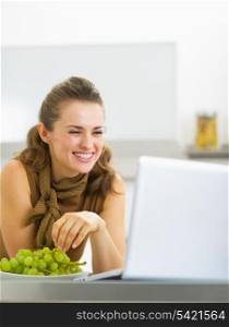 Smiling young woman eating grape and using laptop in kitchen