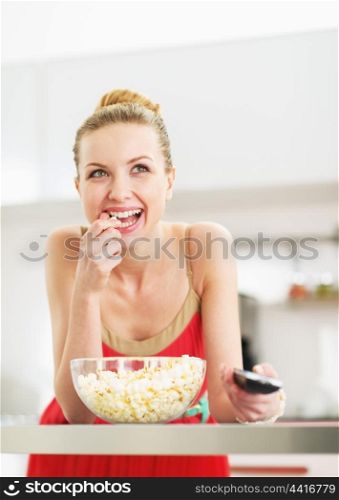 Smiling young woman eating popcorn and watching tv in kitchen