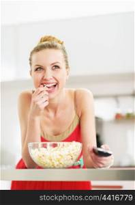 Smiling young woman eating popcorn and watching tv in kitchen
