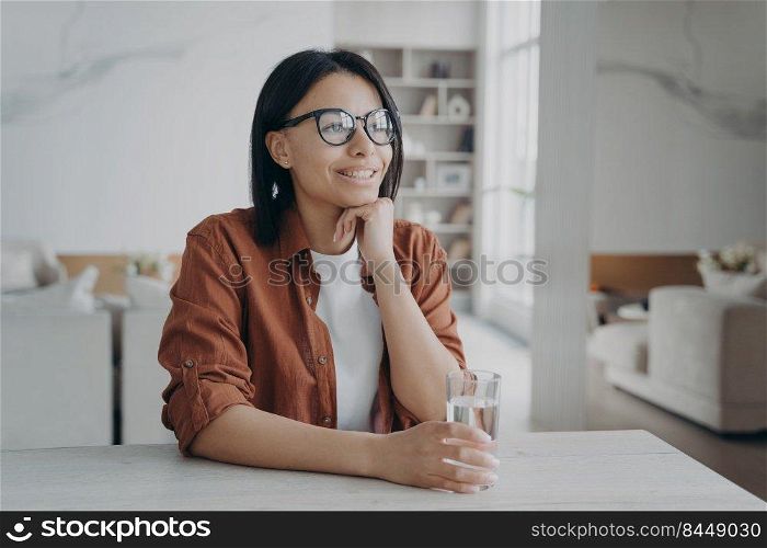 Smiling young woman enjoy purified mineral water holding glass sitting at kitchen at home. Happy female wearing glasses with pure water drink at table. Healthy lifestyle, diet concept.. Smiling woman enjoy purified mineral water holding glass sitting at home. Healthy lifestyle, diet