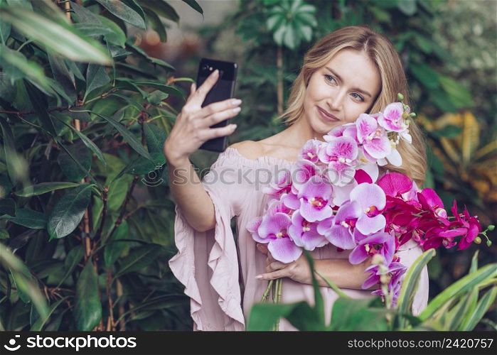 smiling young woman holding beautiful orchid branches hand taking selfie smart phone