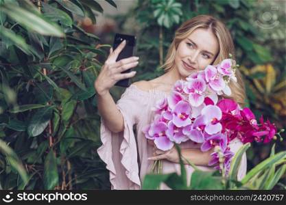 smiling young woman holding beautiful orchid branches hand taking selfie smart phone