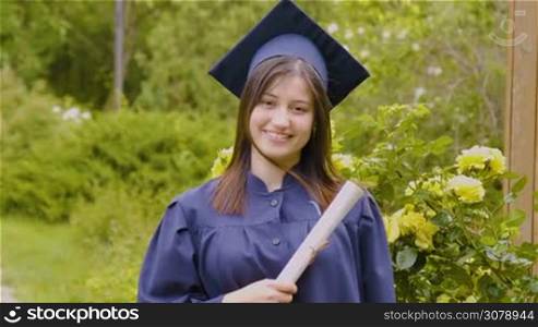 Smiling young woman holding diploma and wearing cap and gown outdoors looking at camera. Graduation concept. Slow motion footage.