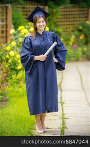 Smiling young woman holding diploma and wearing cap and gown outdoors looking at camera. Graduation concept. . Graduated young woman smiling at camera