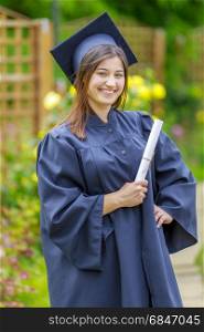 Smiling young woman holding diploma and wearing cap and gown outdoors looking at camera. Graduation concept.