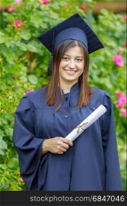 Smiling young woman holding diploma and wearing cap and gown outdoors looking at camera. Graduation concept. . Graduated young woman smiling at camera
