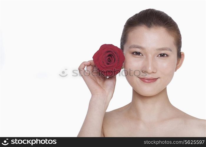 Smiling young woman holding up a red rose next to her ear, studio shot