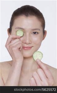 Smiling young woman holding up cucumber slices to put over her eyes, studio shot