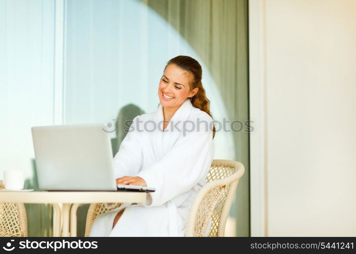 Smiling young woman in bathrobe sitting at table on terrace and using laptop