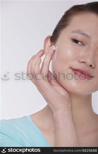 Smiling young woman in blue t-shirt applying cream to her face, studio shot