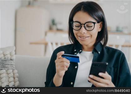 Smiling young woman in glasses holding bank credit card, smartphone in hands, making payment in shopping application, purchasing in online store at home. E-commerce, e-bank, online banking.. Online banking. Woman holding bank credit card, smartphone, making payment in shopping app at home
