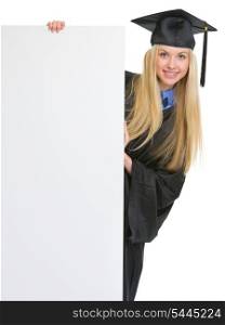 Smiling young woman in graduation gown looking out from blank billboard