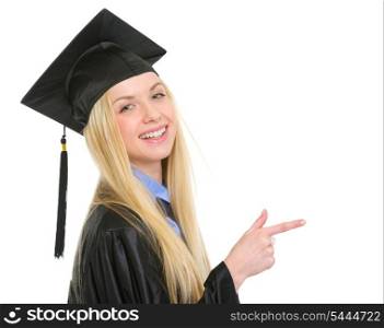 Smiling young woman in graduation gown pointing on copy space