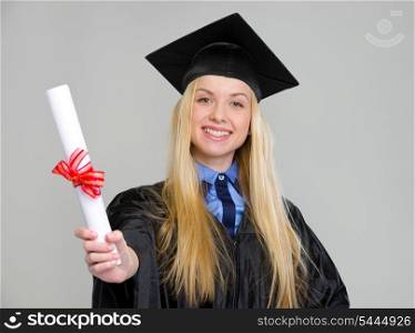 Smiling young woman in graduation gown showing diploma on grey background