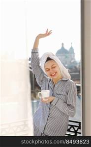 smiling young woman in nightwear and towel on head is holding cup of hot coffee or tea on balcony by the window in the morning. Concept of domestic lifestyle. smiling young woman in nightwear and towel on head is holding cup of hot coffee or tea on balcony by the window in the morning. Concept of domestic lifestyle.