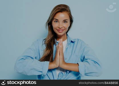 Smiling young woman in oversized shirt and white t-shirt isolated on blue studio background holding hands in prayer Namaste gesture, grateful female looks at camera feel thankful. Smiling young woman in oversized shirt and white t-shirt posing in studio