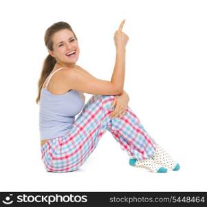 Smiling young woman in pajamas sitting on floor and pointing on copy space