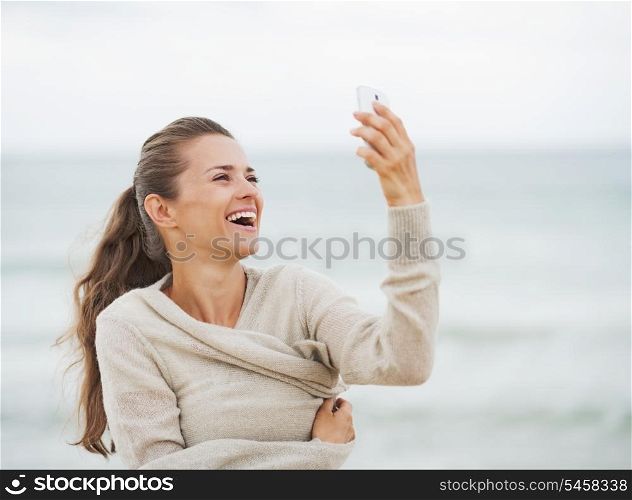 Smiling young woman in sweater on beach taking self photo using cell phone