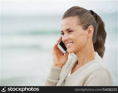 Smiling young woman in sweater on beach talking cell phone