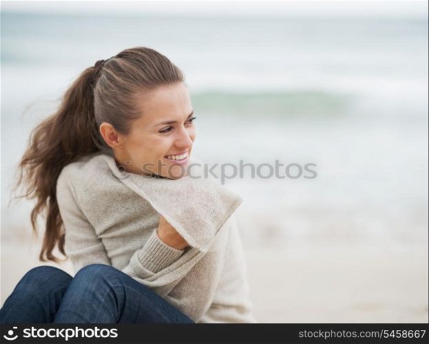 Smiling young woman in sweater while sitting on lonely beach looking on copy space