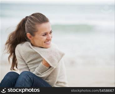 Smiling young woman in sweater while sitting on lonely beach looking on copy space