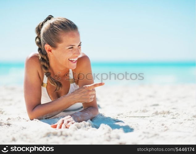 Smiling young woman in swimsuit laying on beach and pointing on copy space