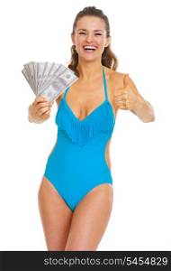 Smiling young woman in swimsuit showing fan of dollars and thumbs up