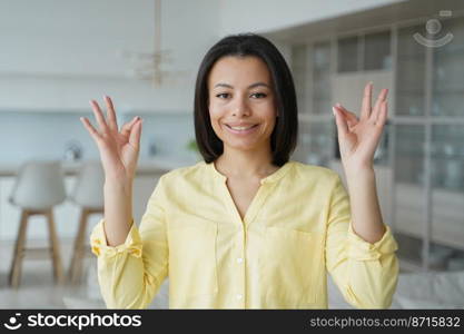 Smiling young woman keeping calm with mudra gesture, practicing yoga breathing exercises at home. Serene female relaxing her mind for stress relief. Healthy lifestyle, wellness, emotional control.. Smiling woman keeping calm with mudra gesture at home. Stress relief, wellness, emotional control