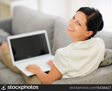 Smiling young woman laying on couch and working on laptop