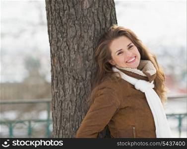 Smiling young woman leaning against tree in winter park