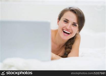 Smiling young woman looking out from laptop while laying in bed