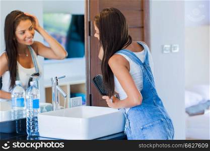 Smiling young woman looking to mirror and brushing hair with comb at home bathroom. Woman brushing hair