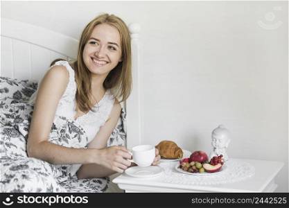 smiling young woman lying bed holding coffee cup with breakfast side table