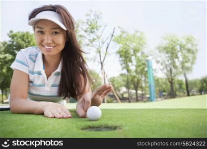 Smiling young woman lying down in a golf course getting ready to flick the ball into the hole