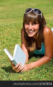 Smiling young woman lying down on grass with book in summer park